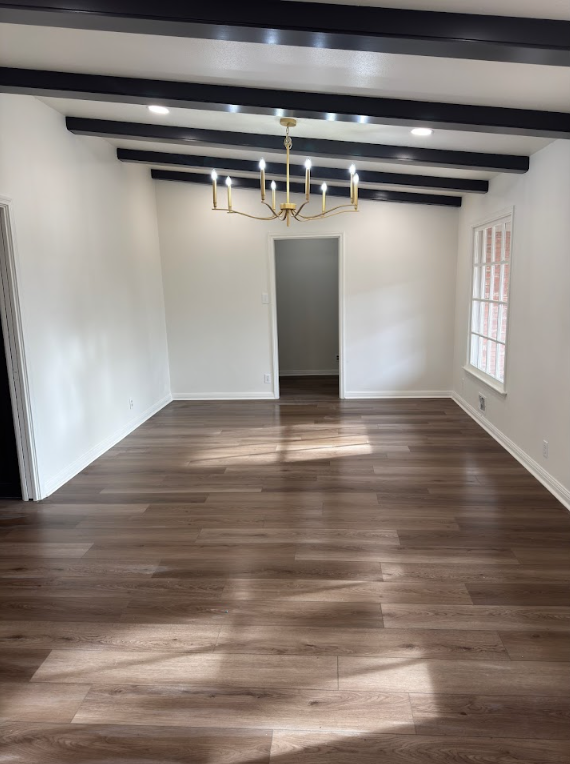 Empty room with wood floors, white walls, and black ceiling beams. A gold chandelier hangs in the center.