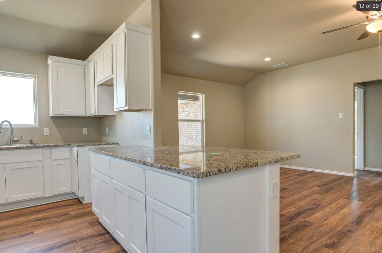 Bright modern kitchen with white cabinets, granite island, and wood flooring leading into an open room
