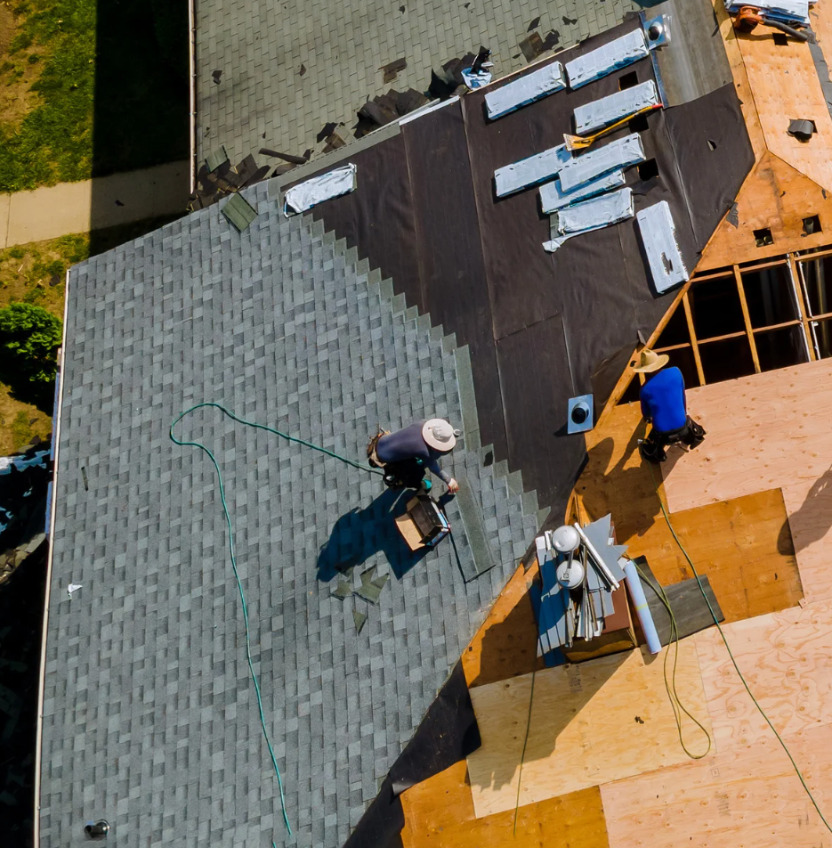 Roofers working on a partially shingled roof, some of the plywood is visible. Sunny day.