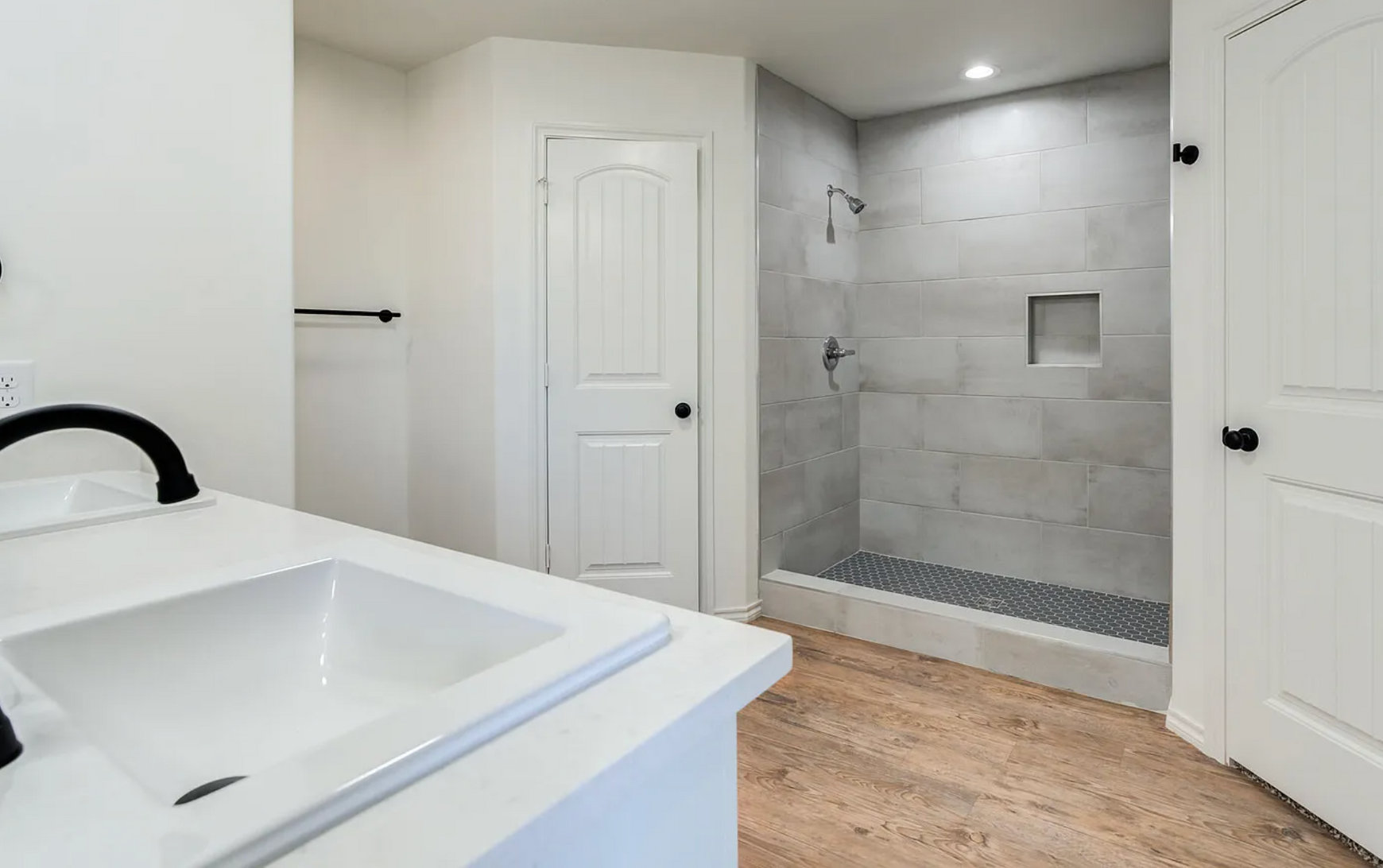 Bathroom with white walls, a sink, shower, wooden floor, and a closed door.