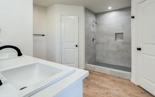 Bathroom with white walls, a sink, shower, wooden floor, and a closed door.