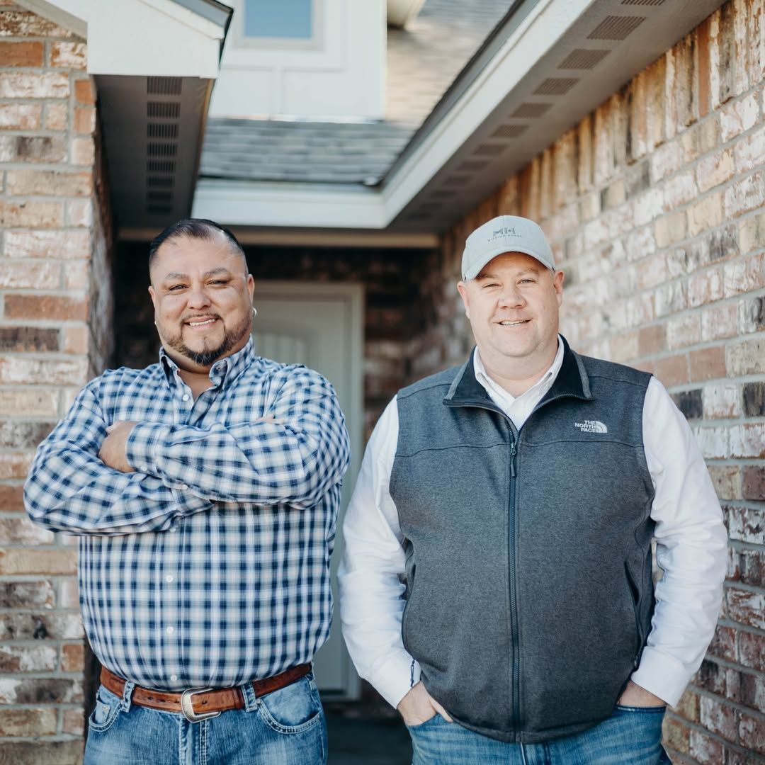 Two men standing in front of a brick building. One with arms crossed, other with hands in pockets.