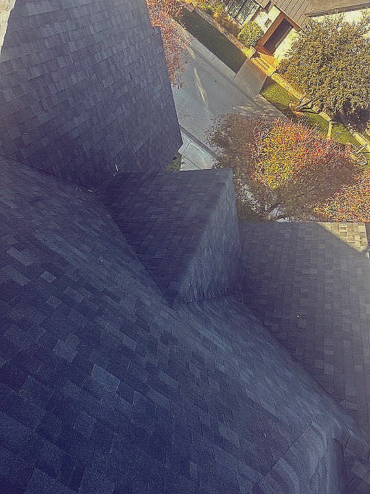 Dark shingled roof with a view of a street, trees, and houses.