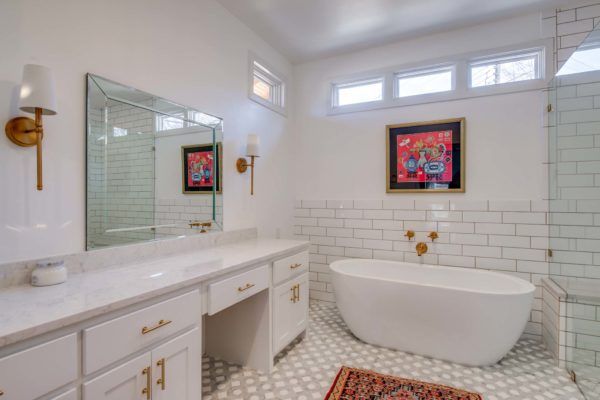 White bathroom with a freestanding tub, vanity, and large mirror. Accented with gold fixtures and art.