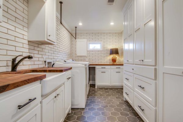 White laundry room with brick-like backsplash, white cabinets, wooden countertop and dark hexagonal floor tiles.