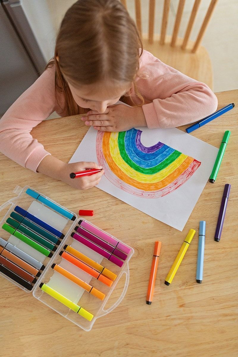 A little girl is sitting at a table drawing a rainbow with markers.