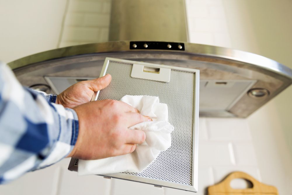 Person cleaning a metal range hood filter with a white cloth in a kitchen.