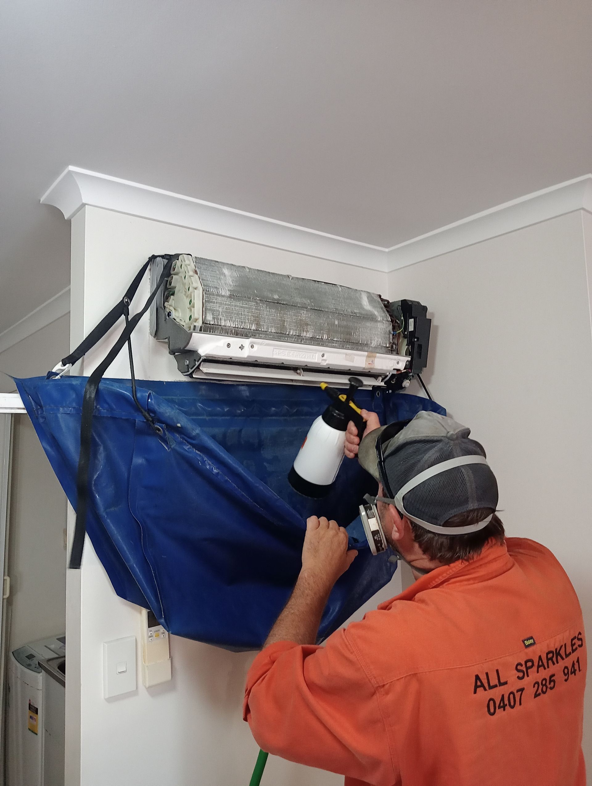 Person cleaning an air conditioner unit with a spray bottle, using a blue cover to catch debris.