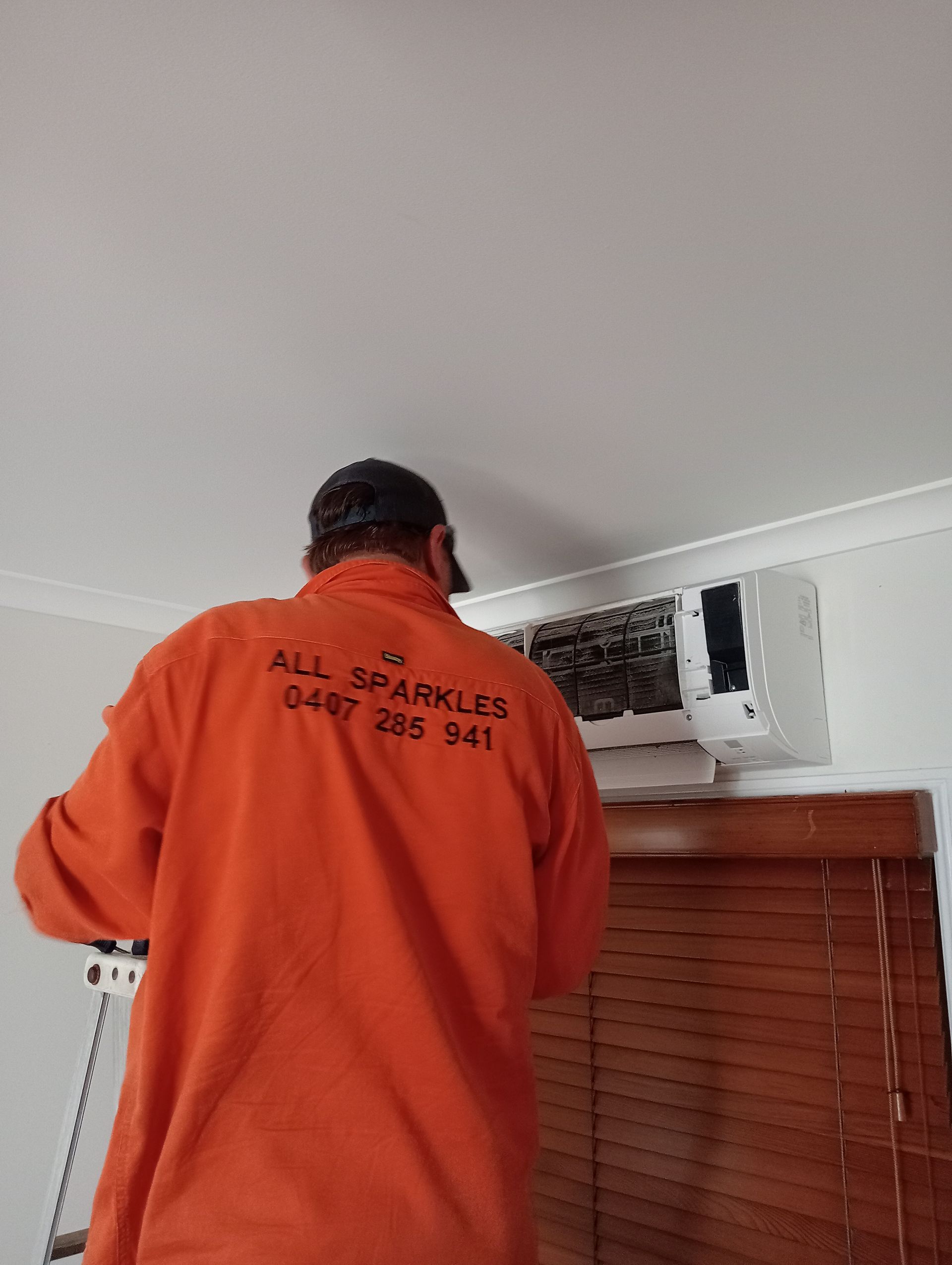 A person in orange shirt working on an air conditioner mounted above a window.
