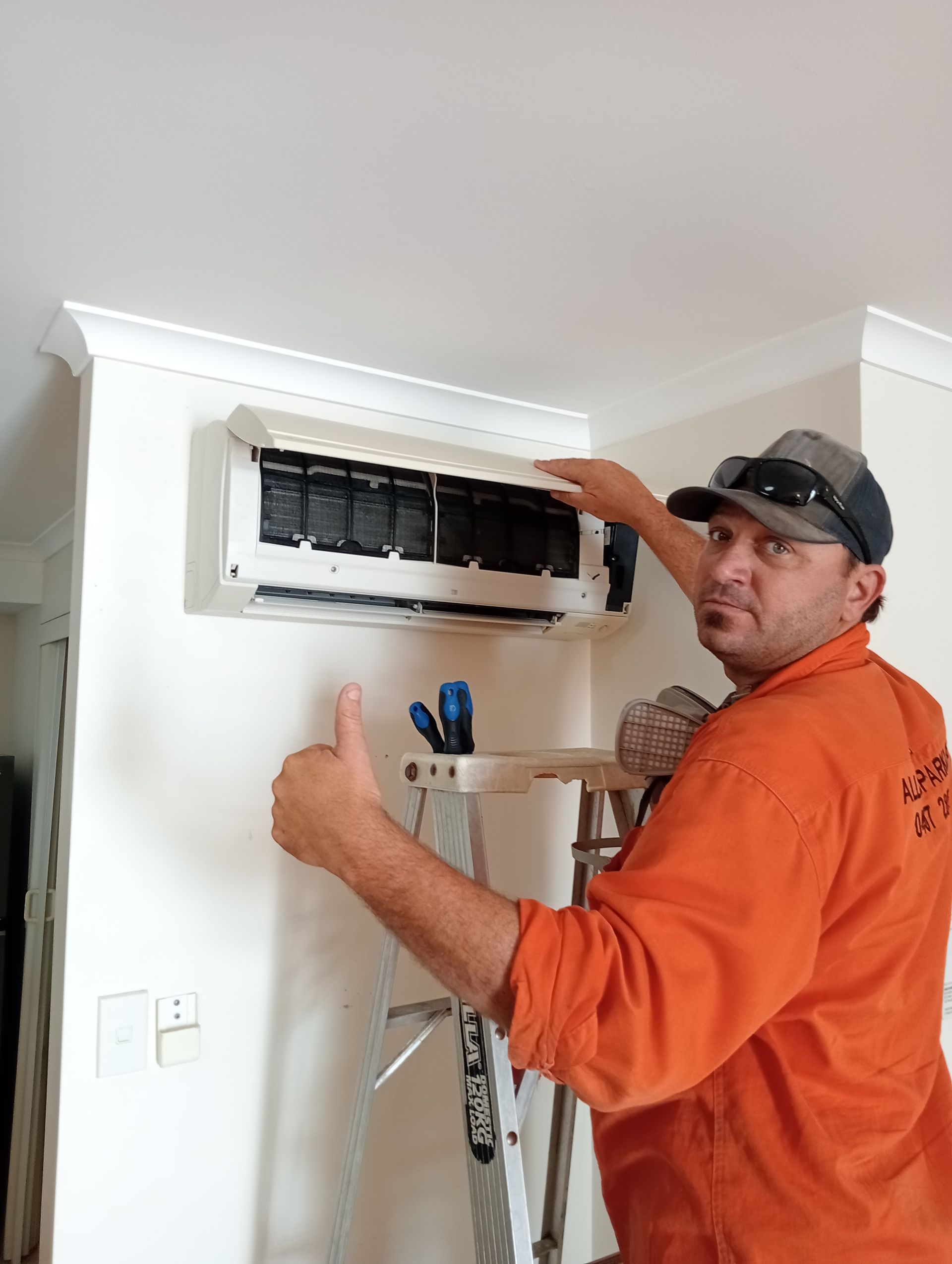 A man in an orange work shirt gives a thumbs-up next to an air conditioner he is working on.