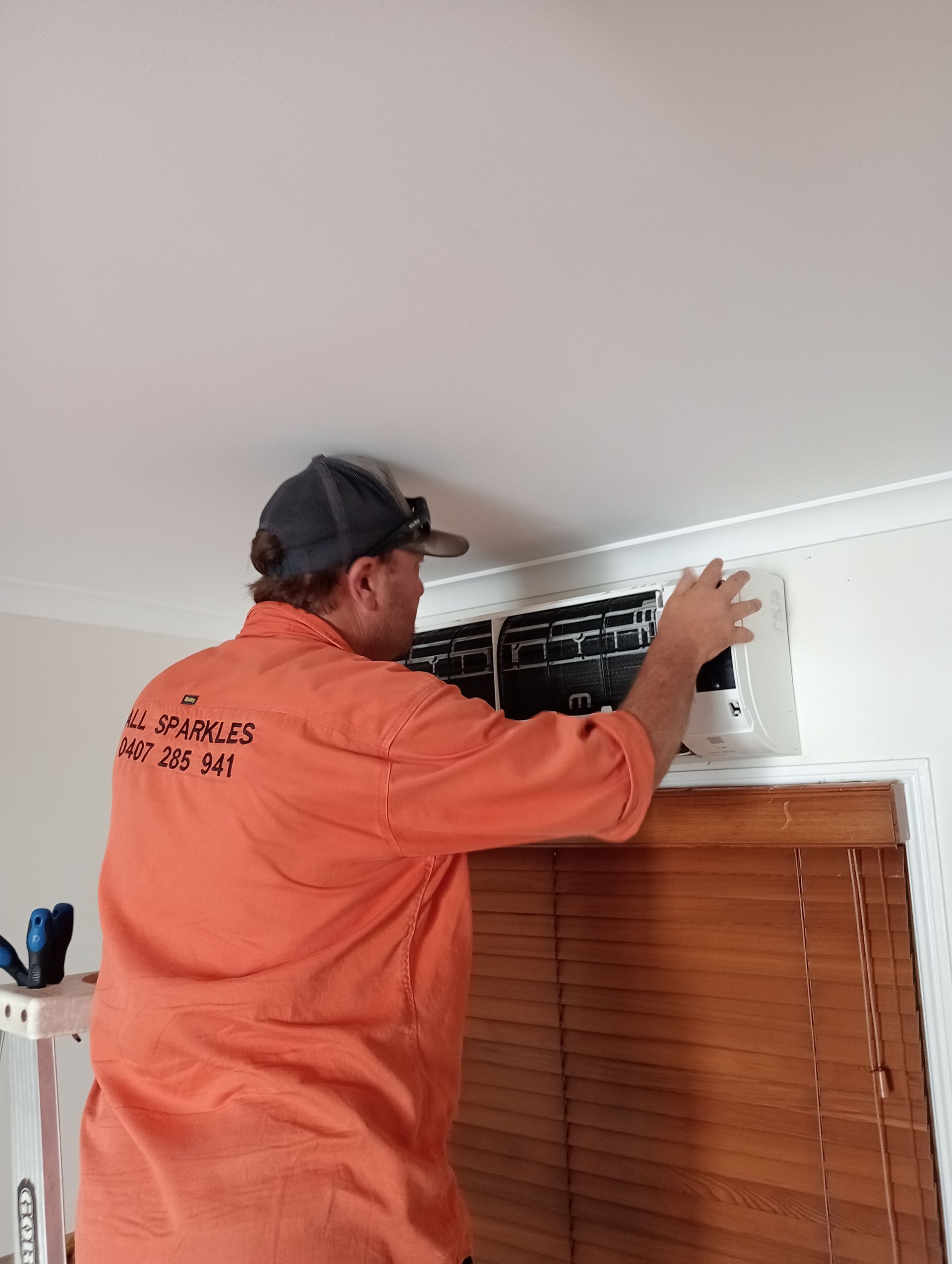 Man in Orange Shirt Installs a Vent on a Wall Above Wooden Blinds — All Sparkles Cleaning Technology In Ayr, QLD