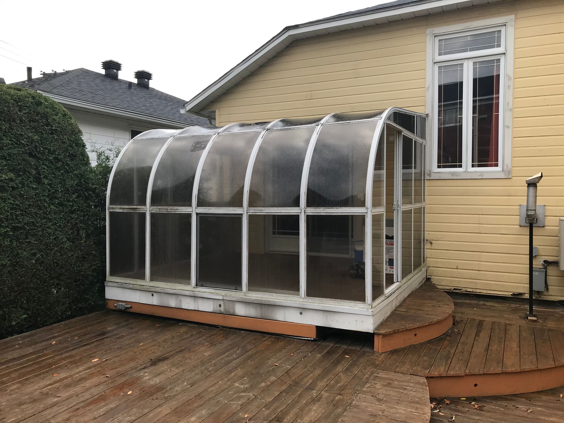 Une serre est posée sur une terrasse en bois devant une maison jaune.
