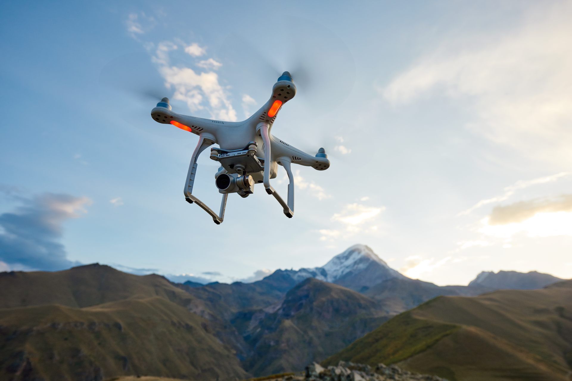 Drone flying over mountains at sunset, with cloudy sky and rugged peaks in the background