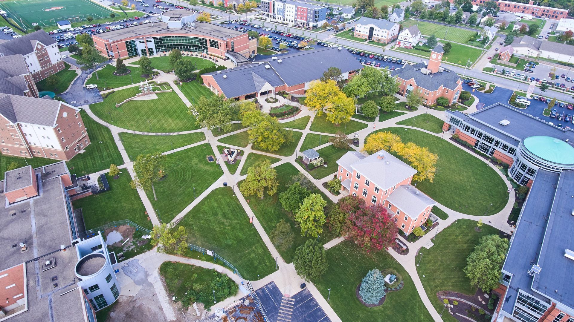 Aerial view of a landscaped park with winding paths, green lawns, trees, and surrounding buildings.