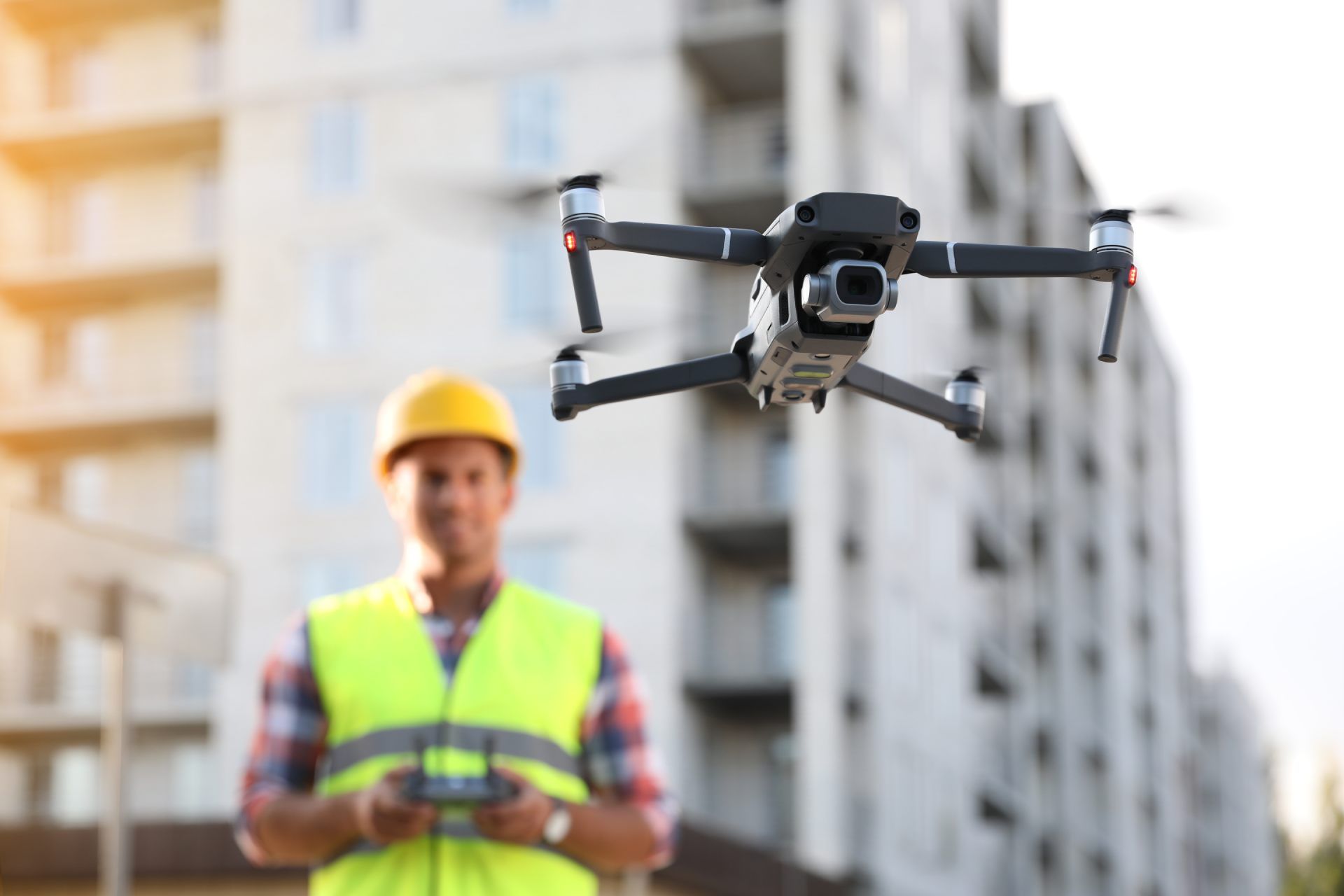 A person operates a drone remote controller during sunset, with a flying drone in the blurred background.