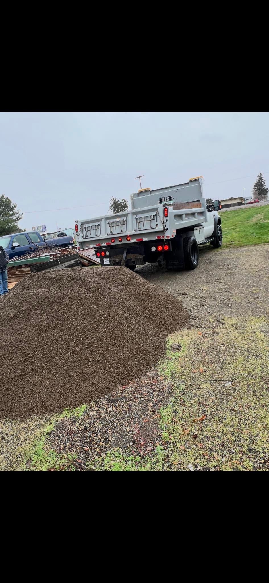 A white dump truck is parked next to a large pile of dark brown gravel on a cloudy day.