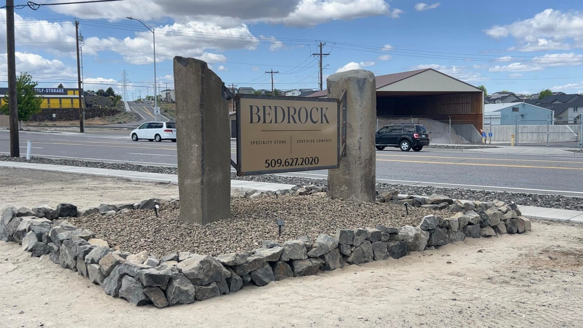 Bedrock sign with two stone pillars, gravel base, and dark metal sign. Buildings and cars in background.