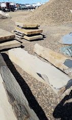 Stack of flagstones and large stones on a gravel lot near a pile of gravel and trucks.