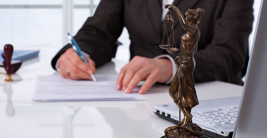 A woman is sitting at a desk writing on a piece of paper next to a statue of justice.