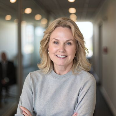 Blonde woman with crossed arms, smiles at the camera. She is in a hallway, wearing a gray sweater.