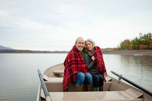 Couple in a rowboat on a lake, wrapped in a red plaid blanket, smiling, overcast sky.