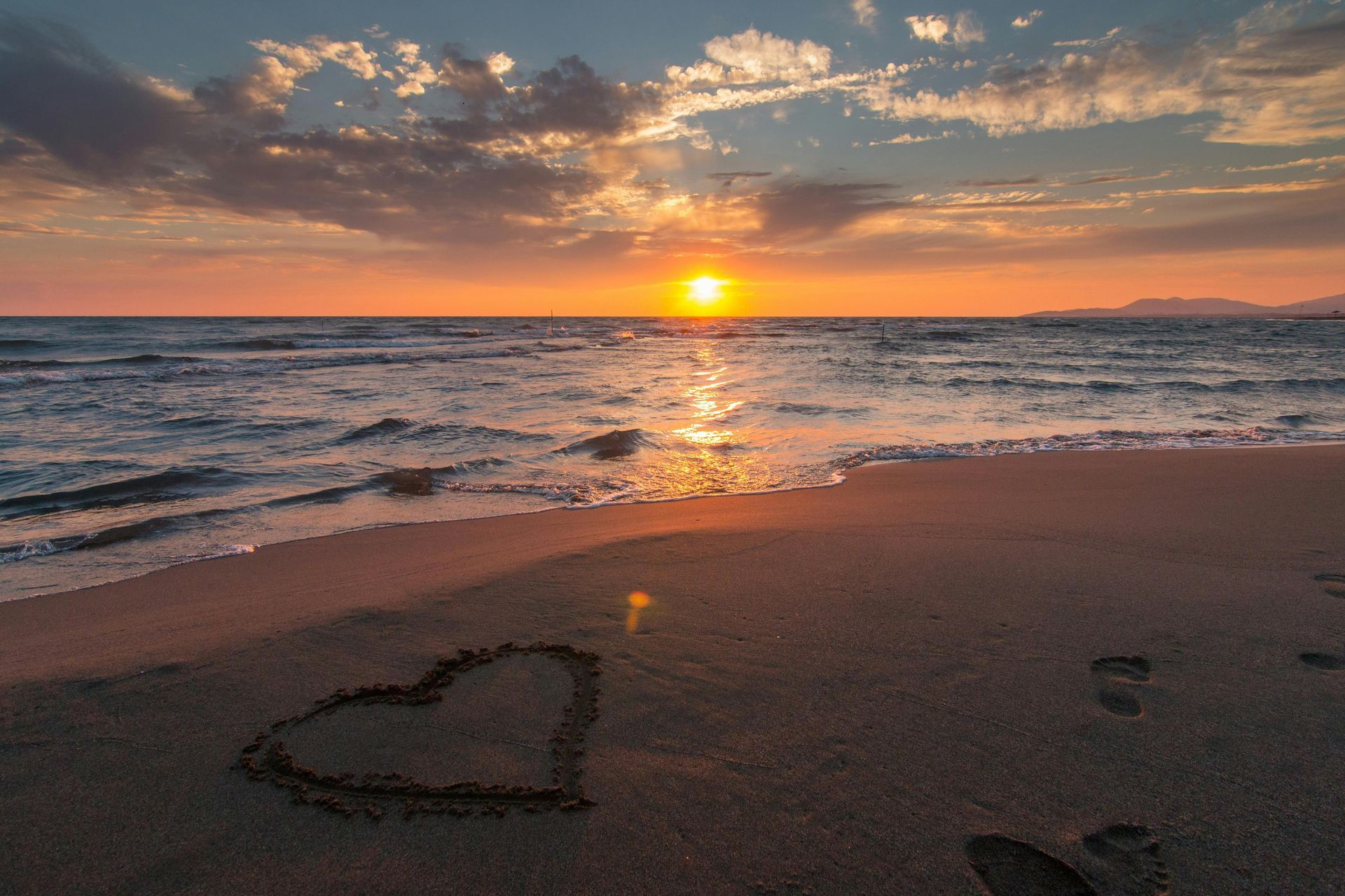 Sunset over ocean; heart drawn in sand on beach.