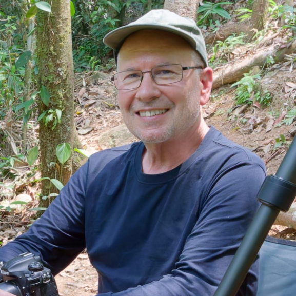 Smiling person in a cap and glasses standing on a forest trail with a bicycle.