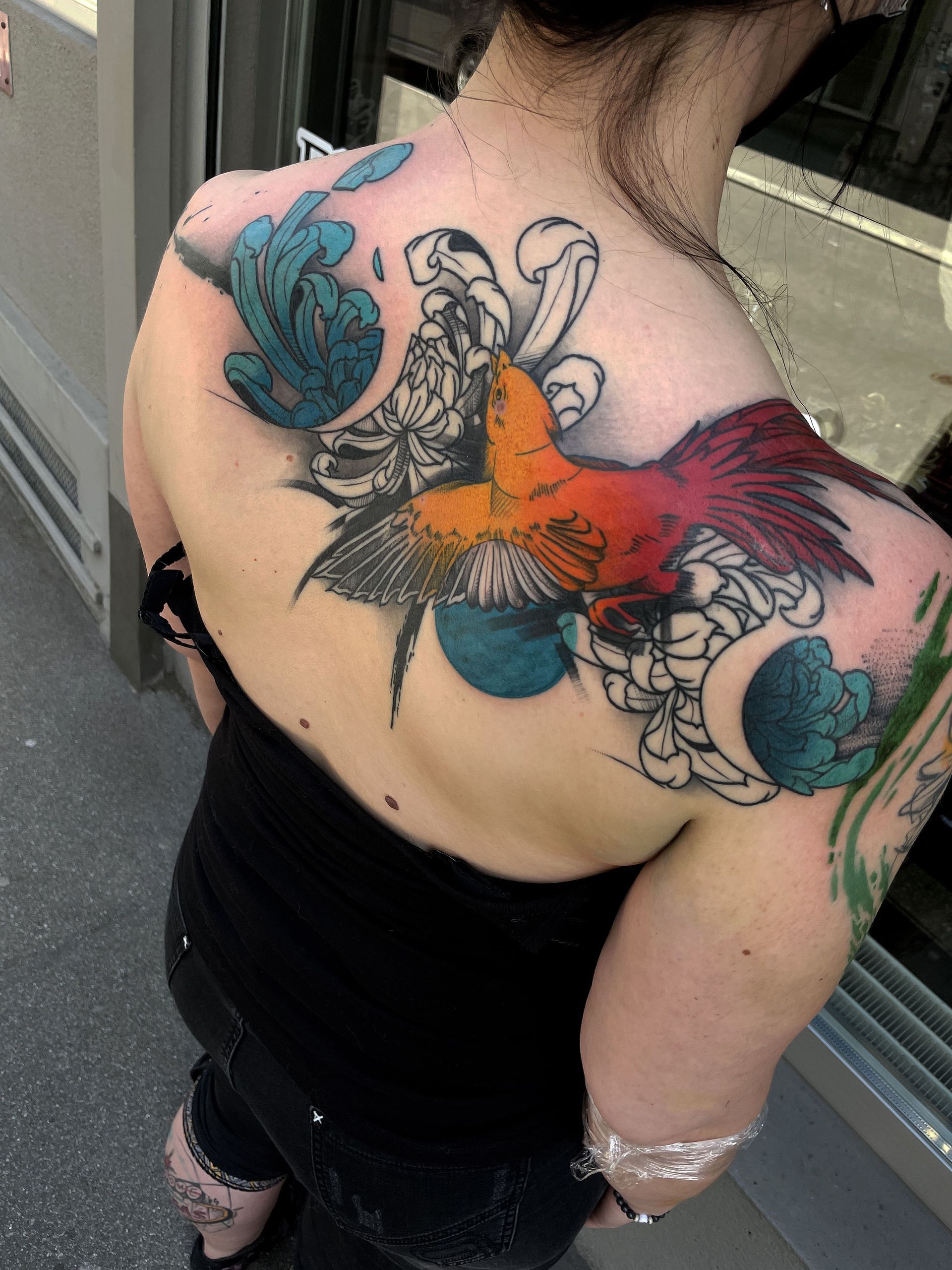 Tätowierung auf dem Rücken einer Person: bunter Vogel, blaue Akzente, Blumenmuster. Schwarzes Oberteil, im Freien.