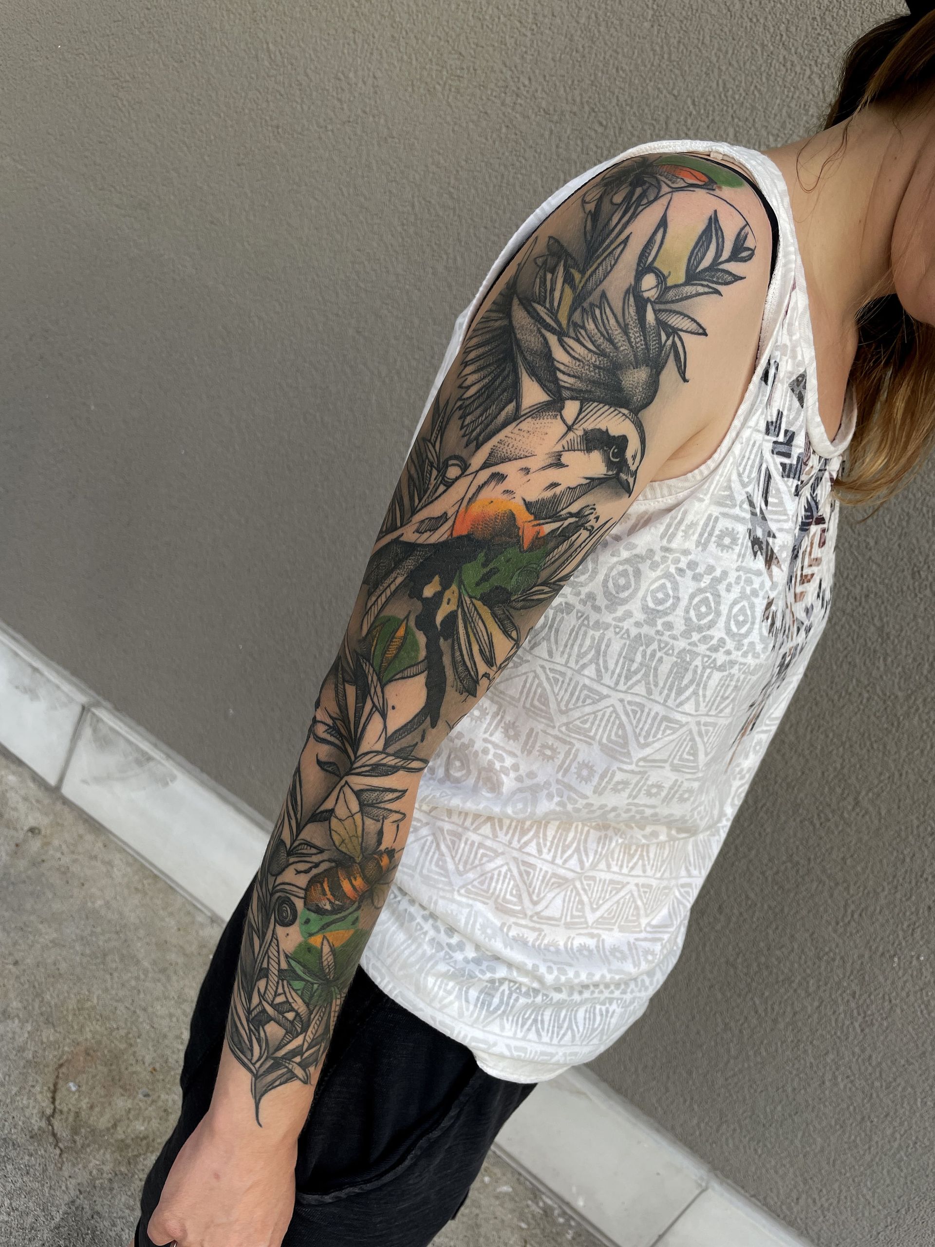 Person mit einem Ganzkörpertattoo auf dem linken Arm, die ein weißes Tanktop trägt und an einer Wand steht.
