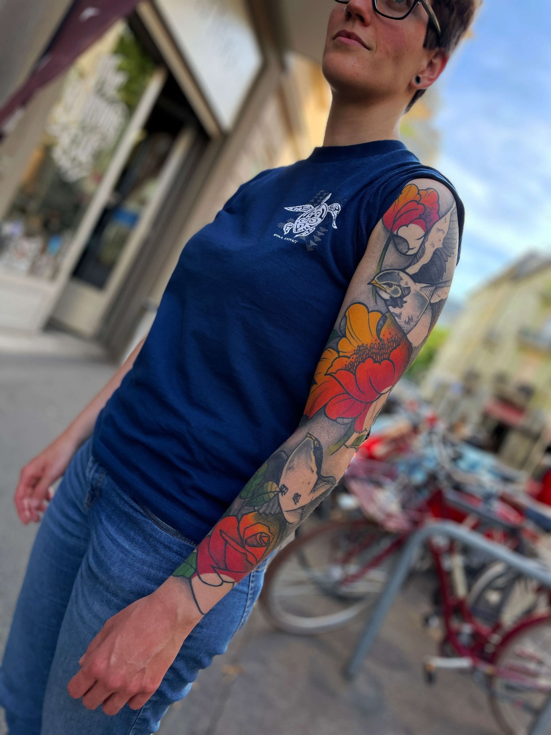 Person mit buntem Blumentattoo am Arm, die ein blaues Hemd und Jeans trägt und draußen steht.