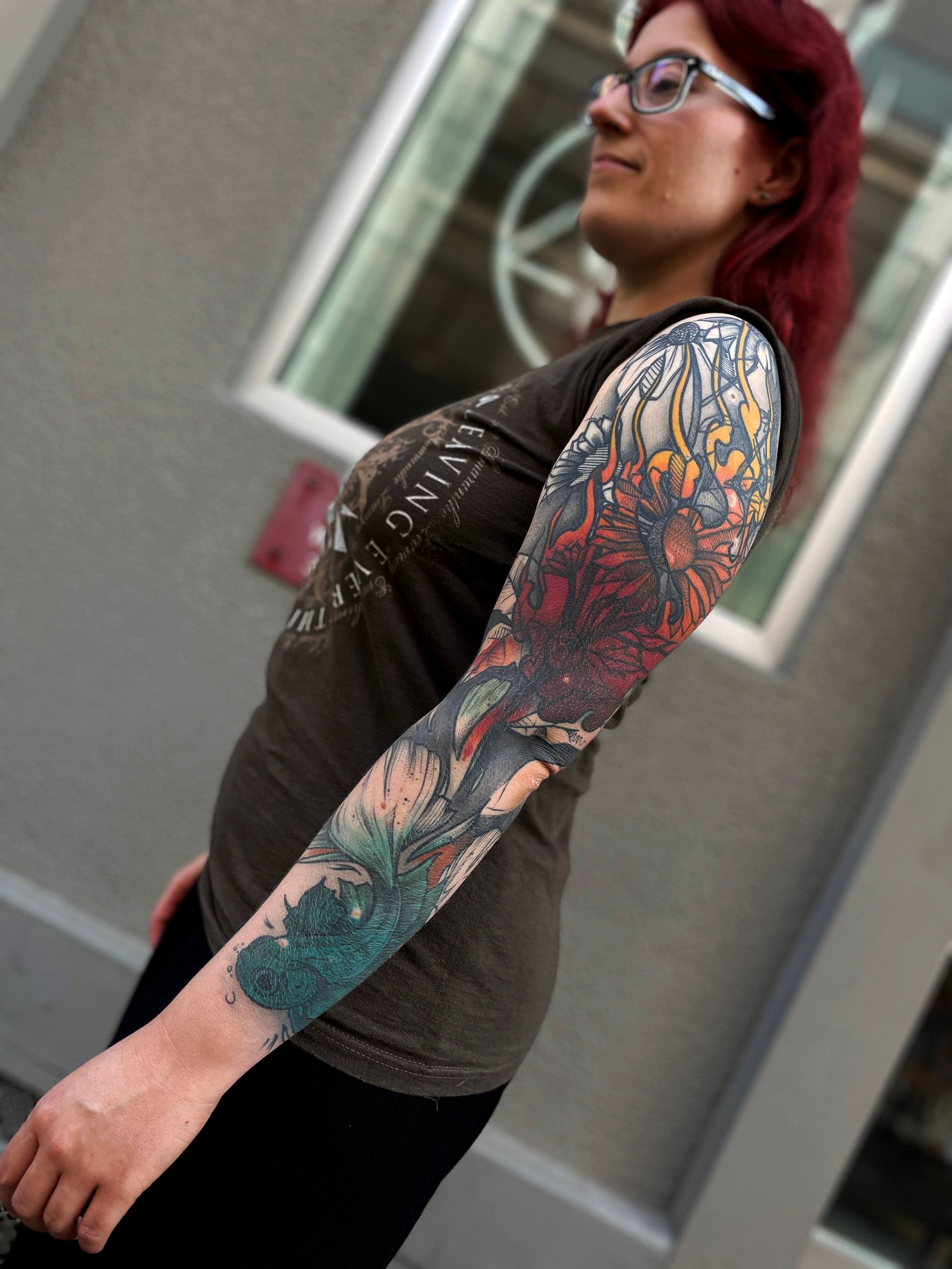 Person mit roten Haaren und Brille, Ganzarm-Tattoo, trägt ein dunkles T-Shirt. Steht draußen in der Nähe eines Fensters.