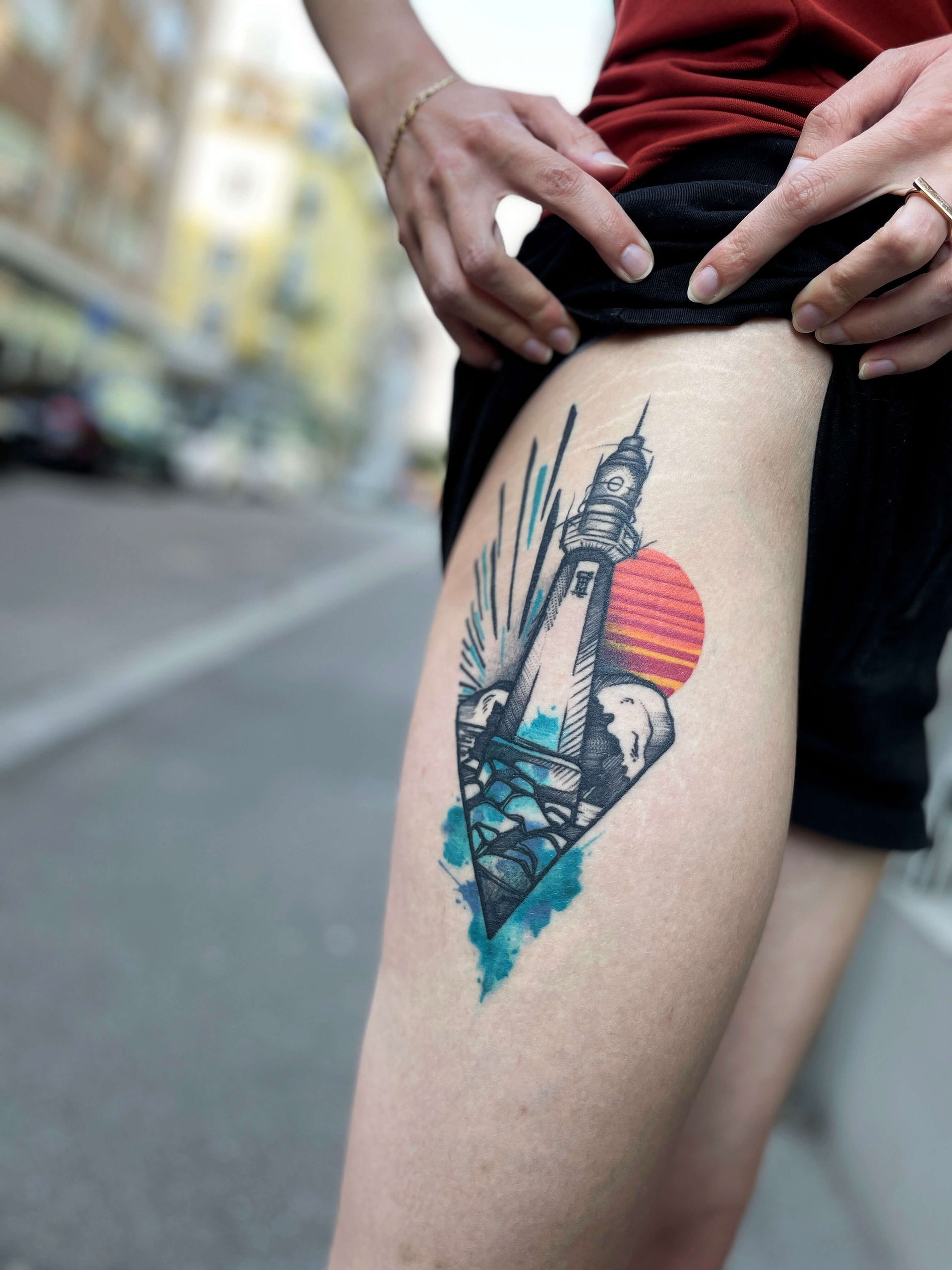 Leuchtturm-Tattoo auf dem Bein mit untergehender Sonne, blauem Wasser und Gischt; Person in Shorts.