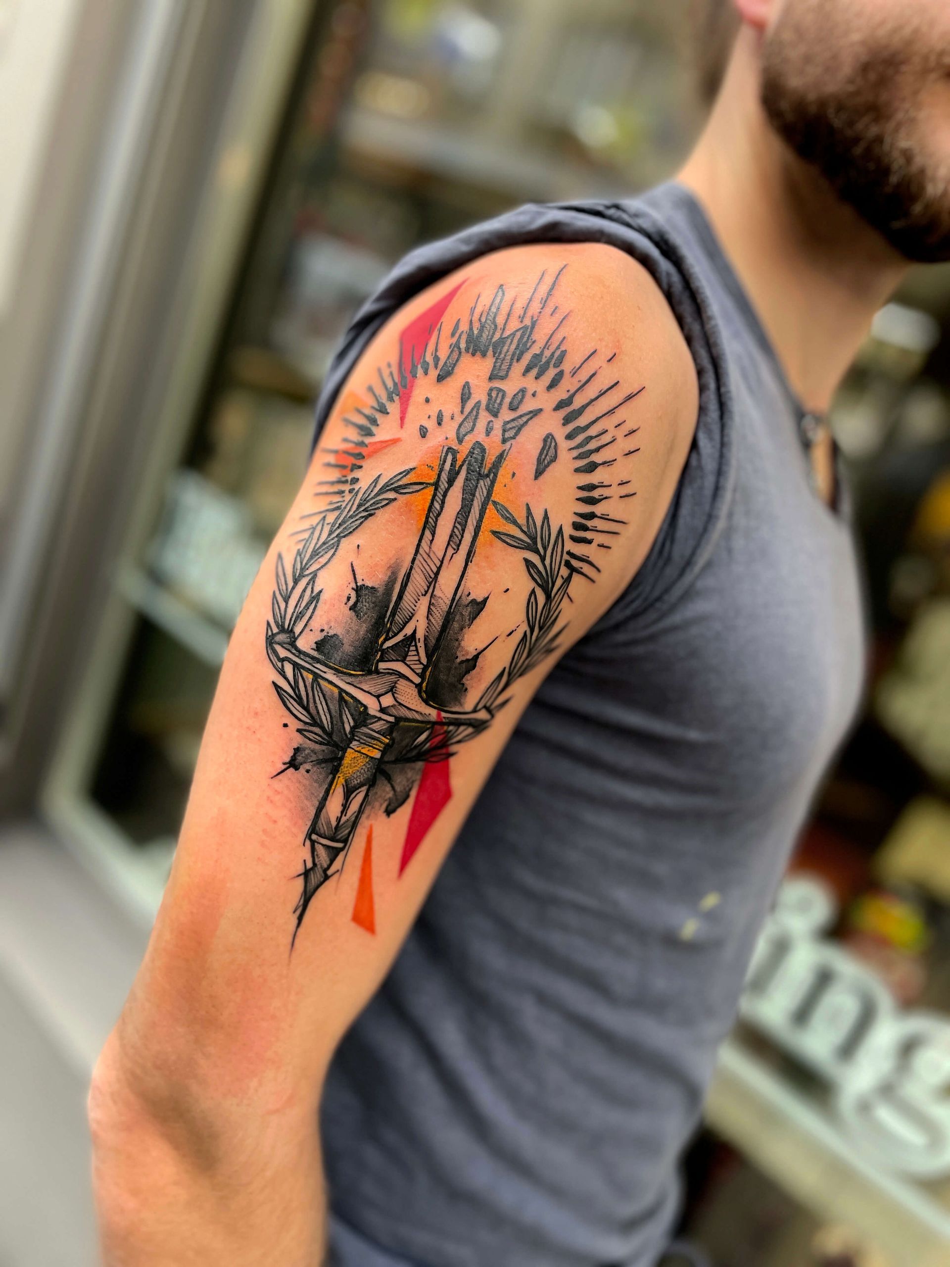 Tätowierung eines Schwertes mit Lorbeerkranz auf dem Oberarm einer Person, mit rot/orangen geometrischen Formen.