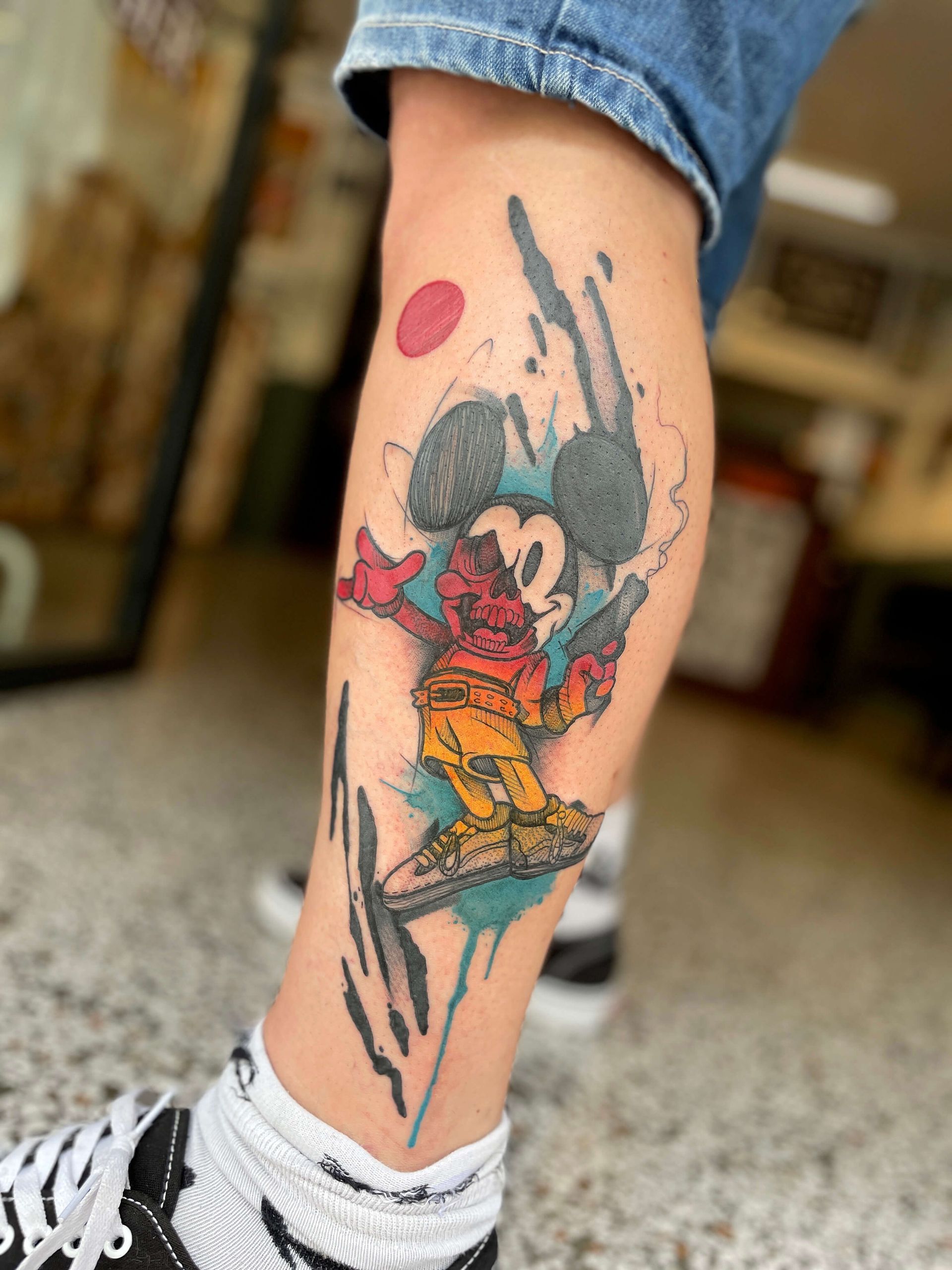Tattoo von Mickey Mouse mit einem bunten, abstrakten Design auf einem Bein.