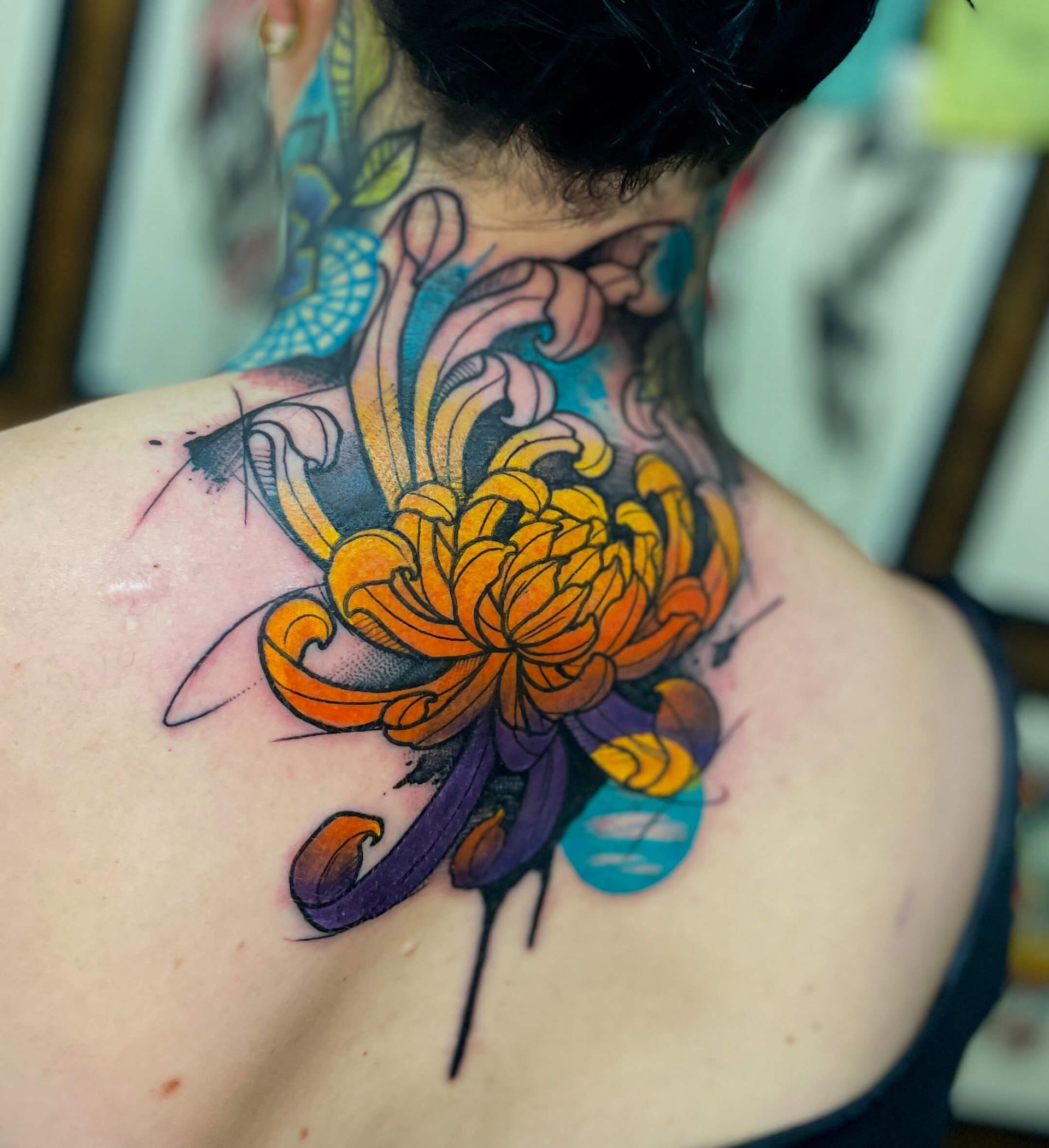 Rückentattoo einer orange-violetten Chrysantheme mit blauen und gelben Akzenten am Hals einer Person.