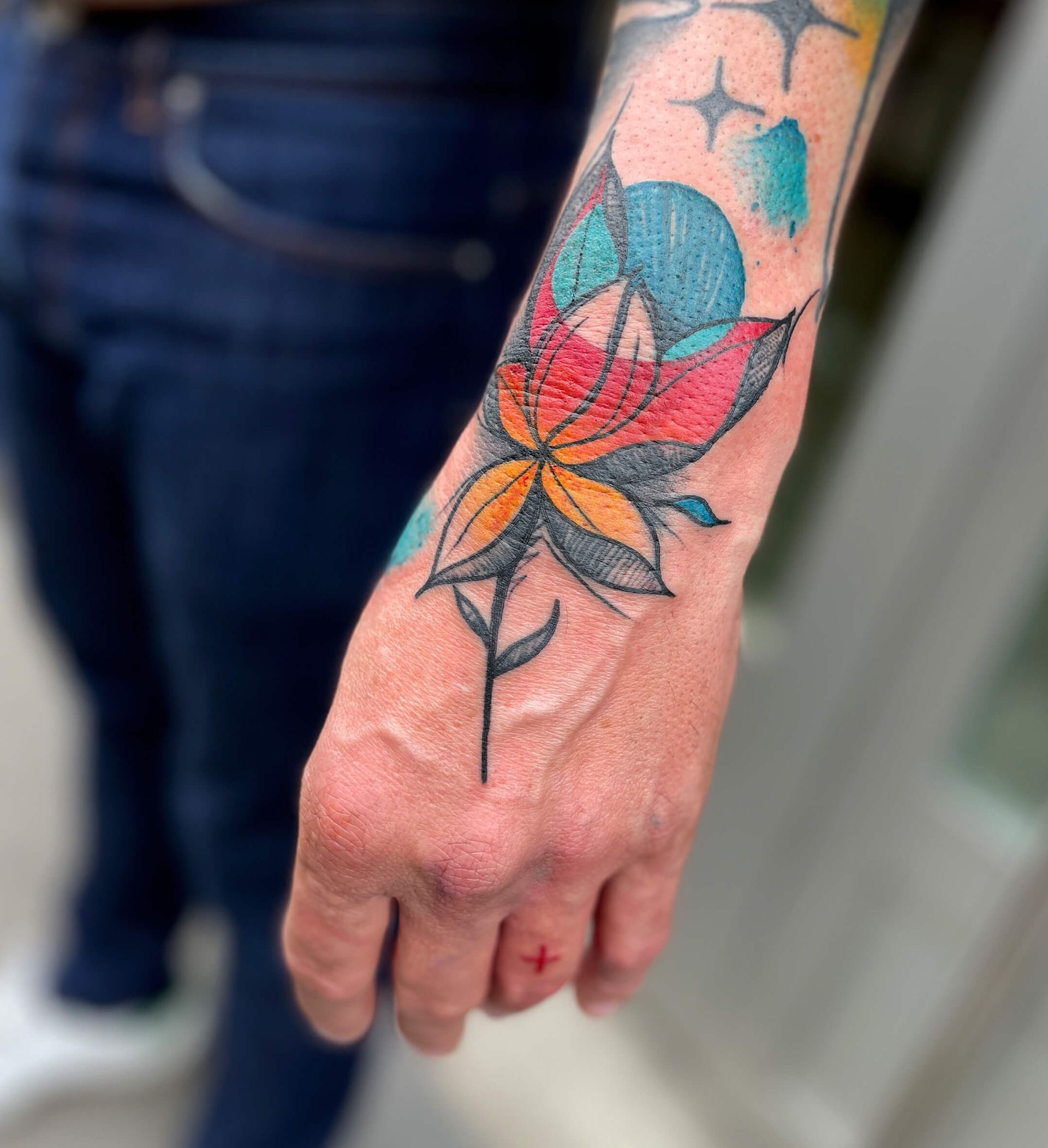 Buntes Blumentattoo am Handgelenk einer Person; blaue, orange, rote, gelbe Blütenblätter, Sterne, dunkler Stiel.