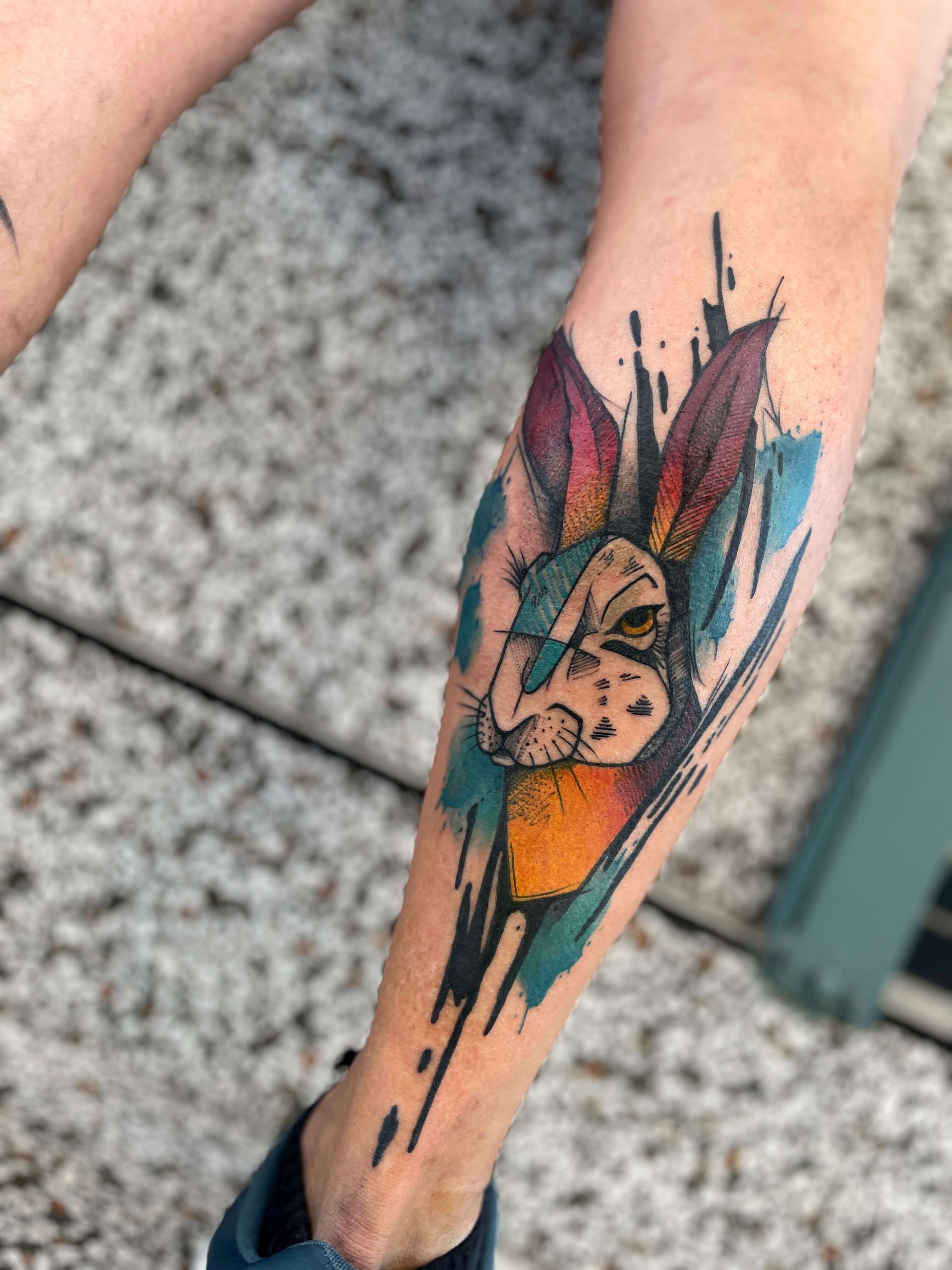 Tätowierung eines Kaninchengesichts mit bunten Pinselstrich-Akzenten auf einer Wade.