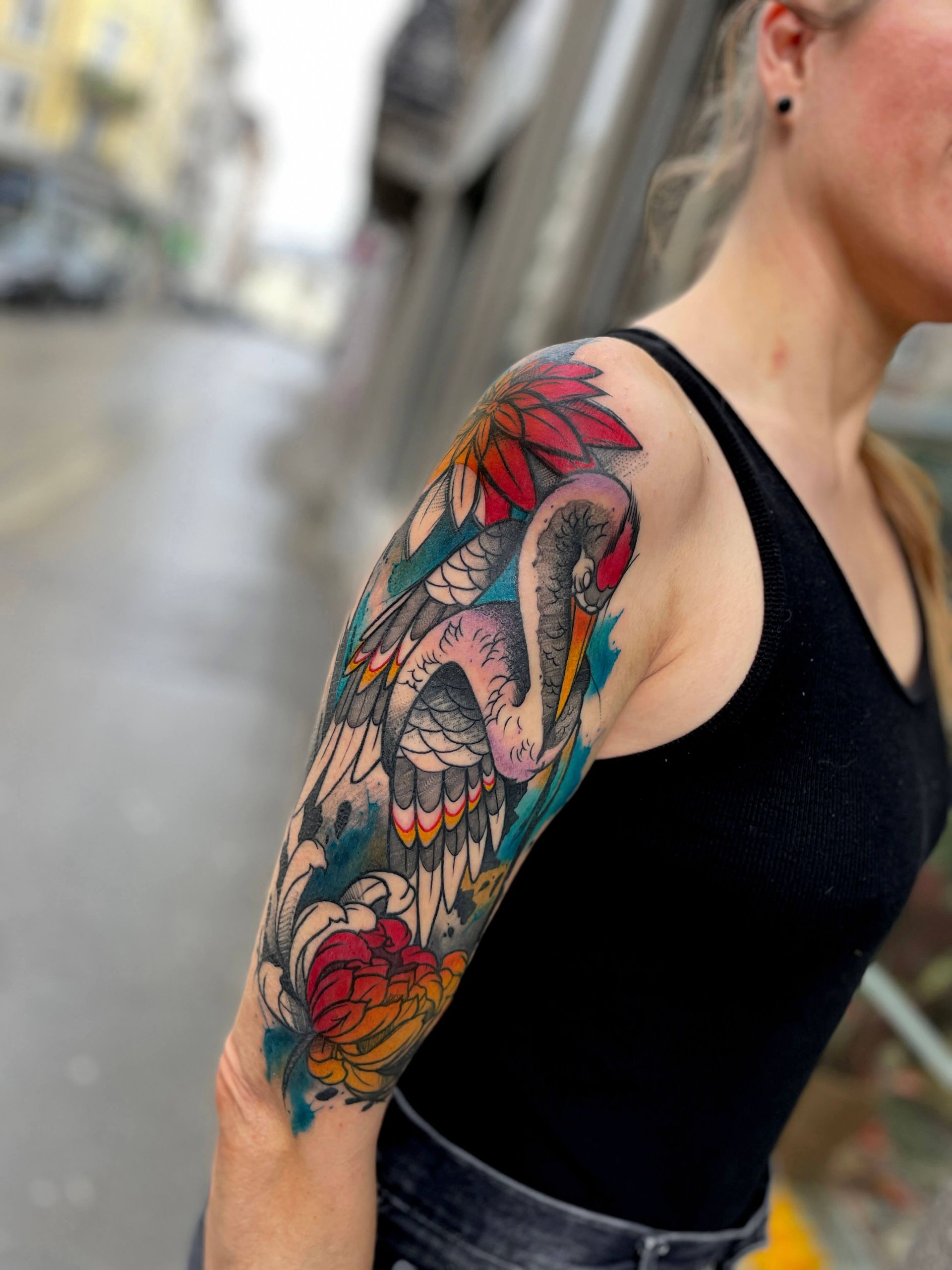Frau mit buntem Armtattoo eines Kranichs und Blumen auf einer Stadtstraße.