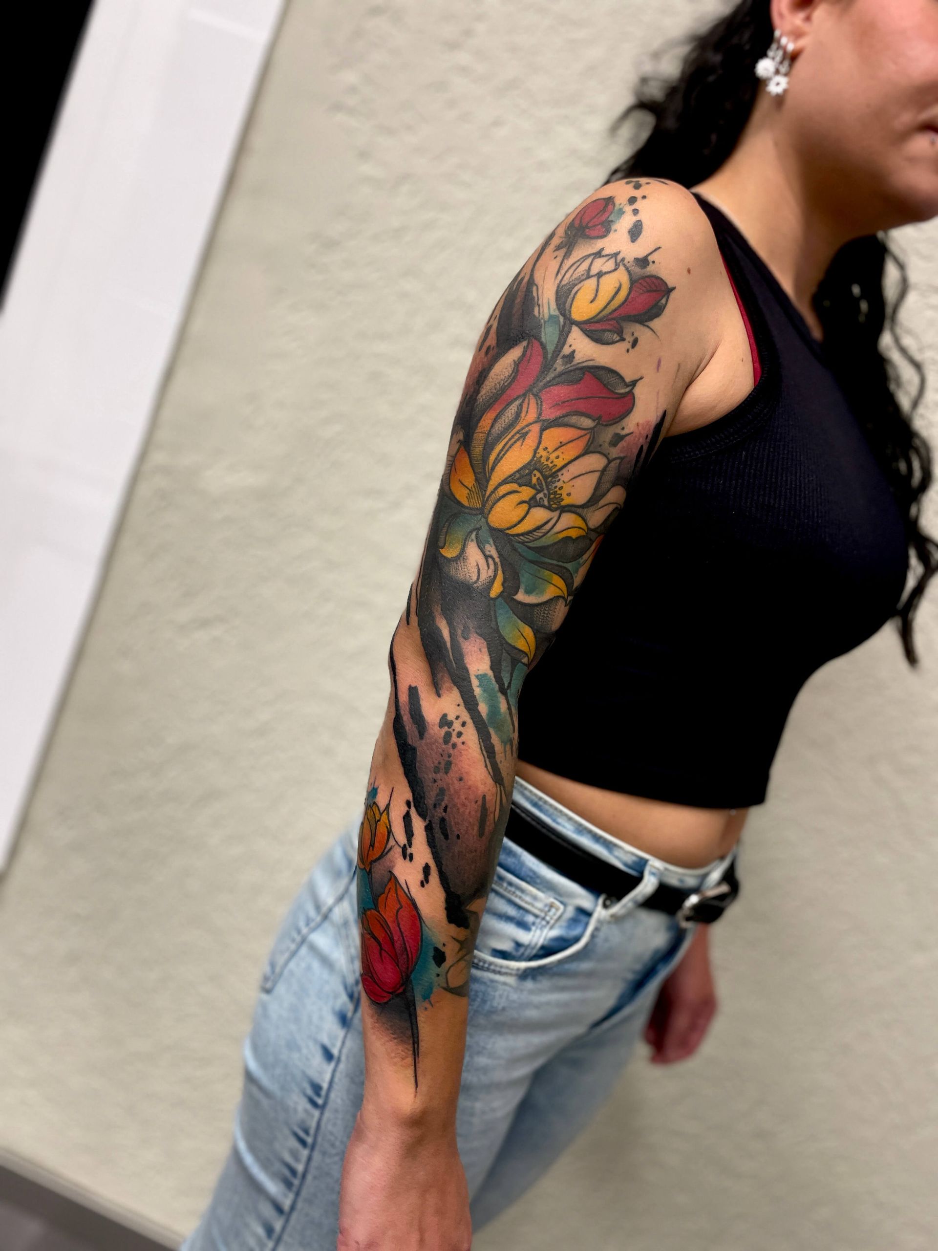 Frau mit buntem Blumentattoo auf dem Arm, trägt schwarzes Tanktop und Jeans.