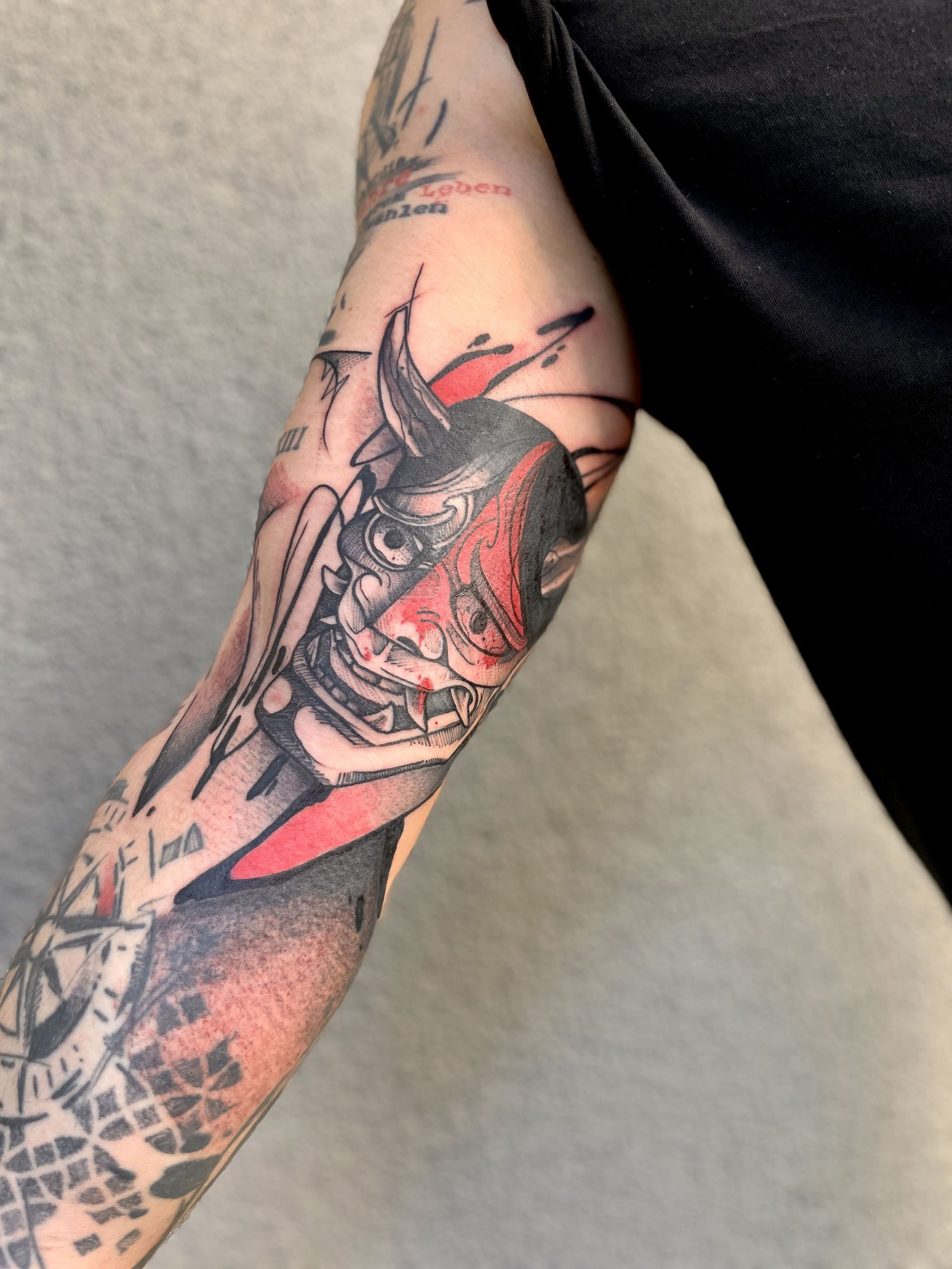Tätowierung einer rot-schwarzen Dämonenmaske auf einem Unterarm, mit schwarzer Schattierung und Windrose.