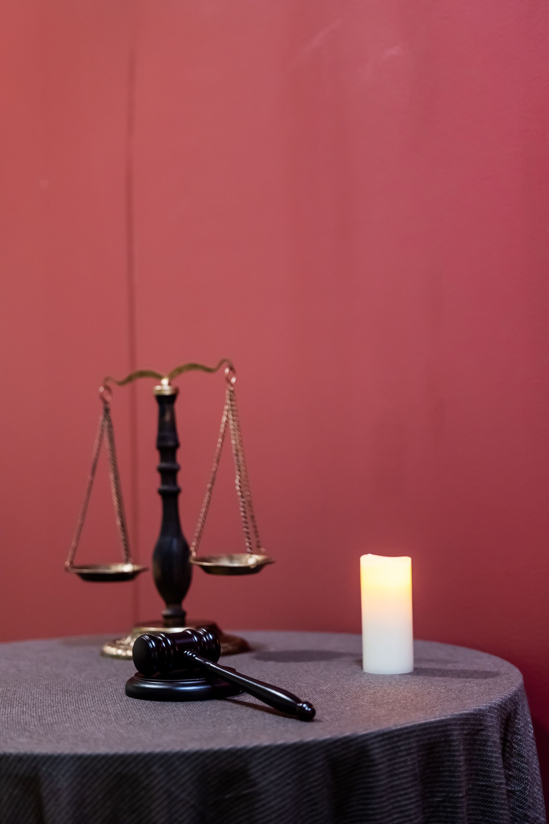 Balanced scales, a gavel, and a lit candle on a dark table against a red background.