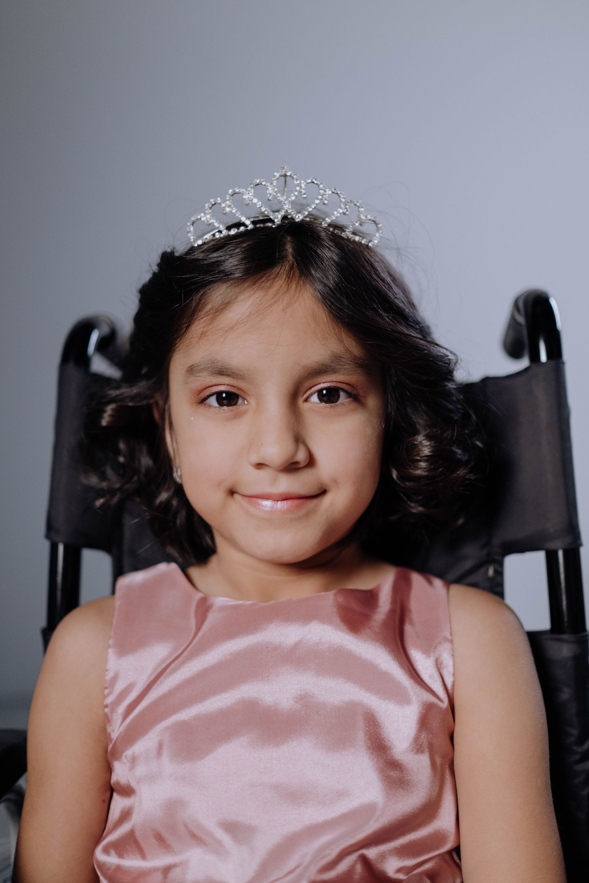 Young child in a pink dress and tiara, smiling while seated in a black chair