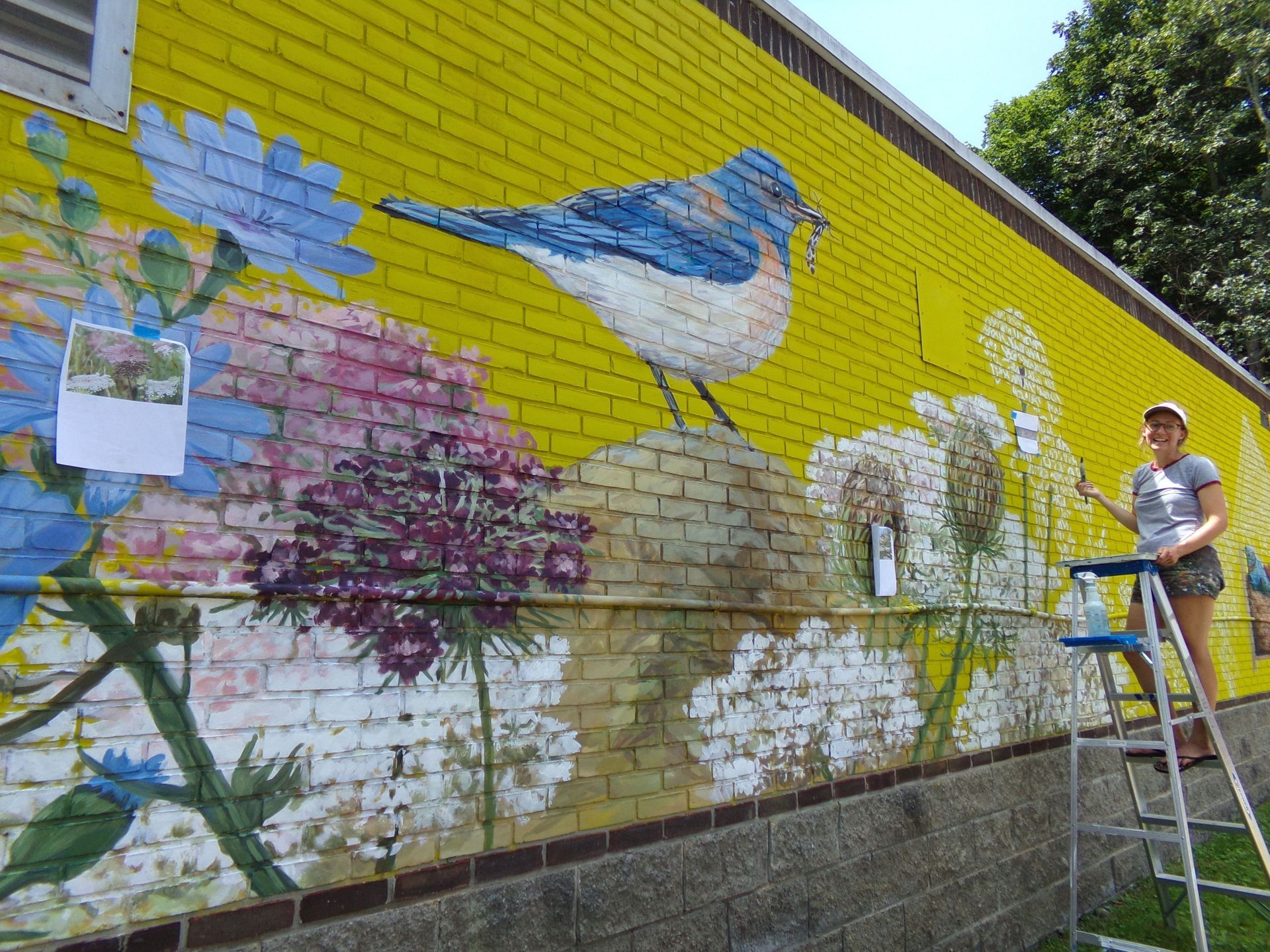 Chloe Smith painting a colorful bird mural on a yellow brick wall beside a ladder