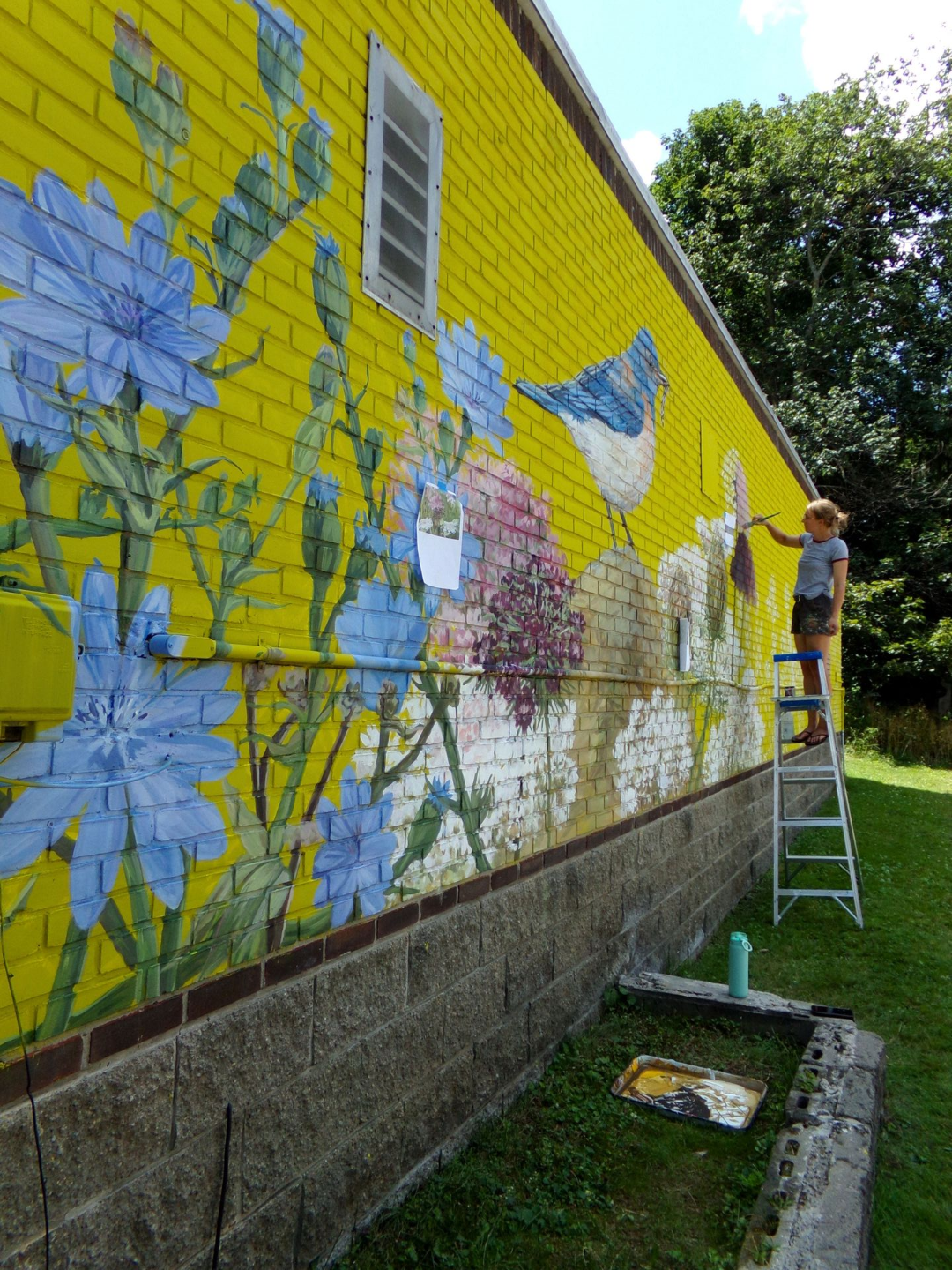 Person painting a yellow brick wall mural with blue flowers from a ladder outdoors