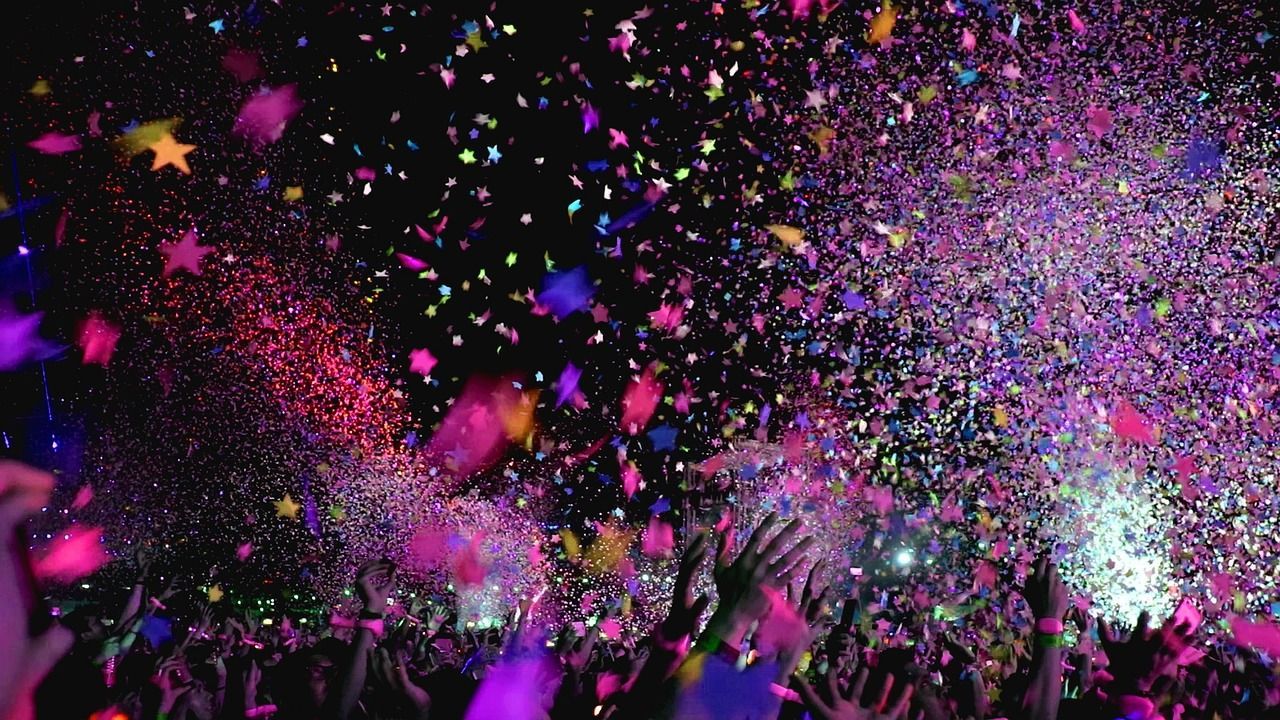 Colorful confetti falling on a crowd during a nighttime celebration, with people raising their hands under a star-shaped light display.
