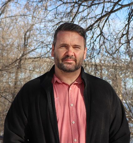 Man in a black jacket and pink shirt standing outdoors with bare trees and a clear sky in the background.