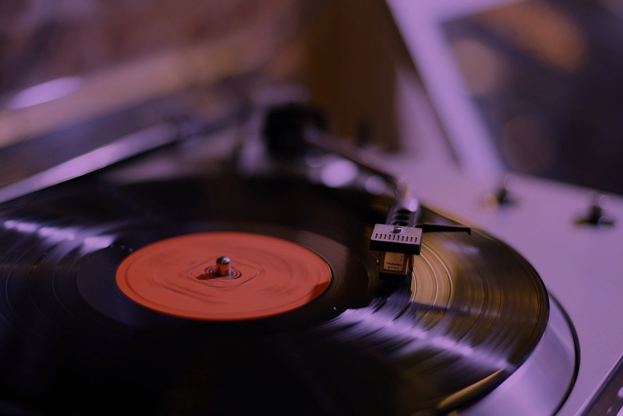 A close-up of a vinyl record spinning on a turntable with a stylus resting on the record, illuminated in soft purple lighting.