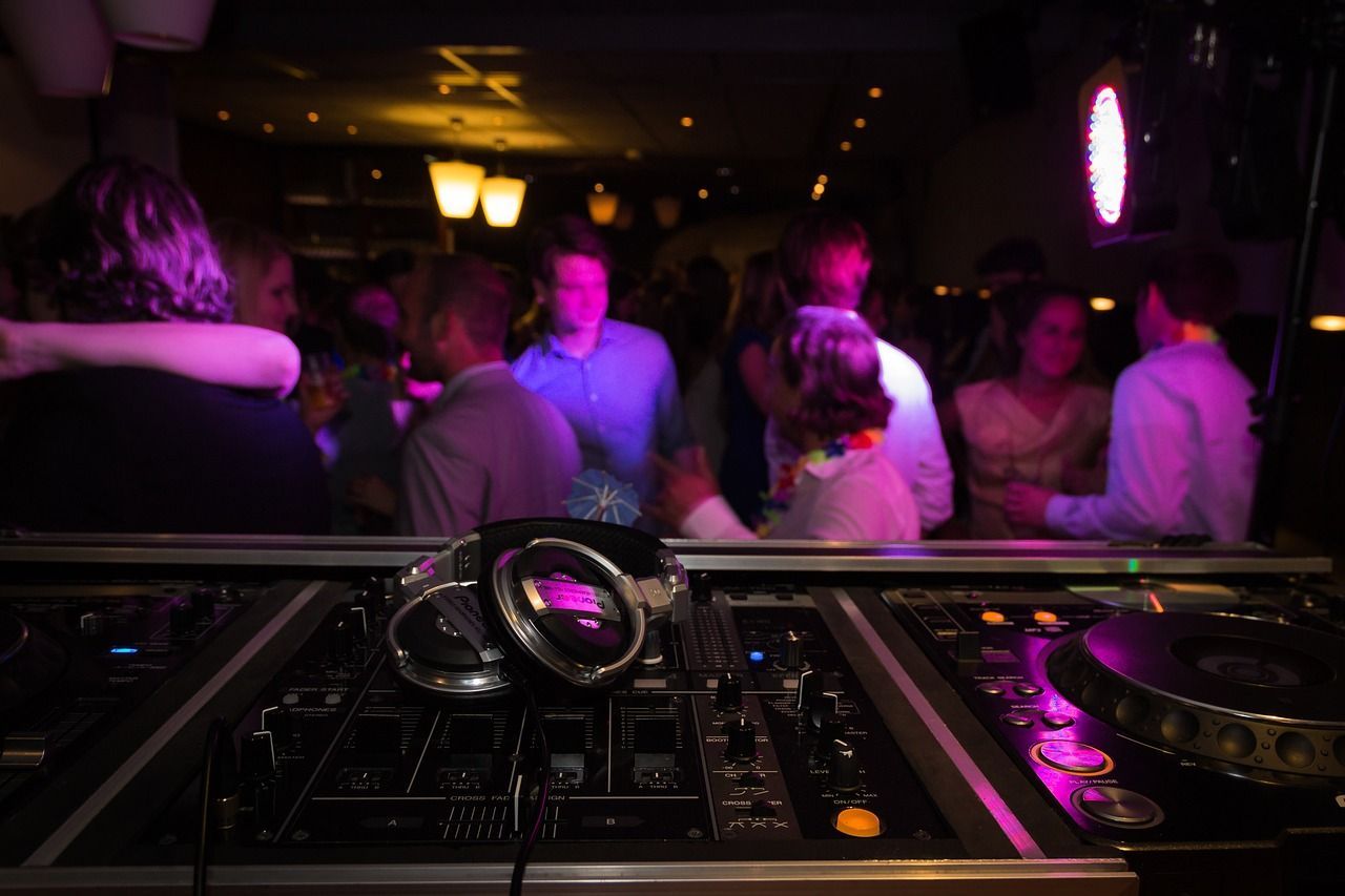 Dj equipment, including headphones and a mixer, in focus with a blurry background of people dancing under dim, colorful lights at a party.