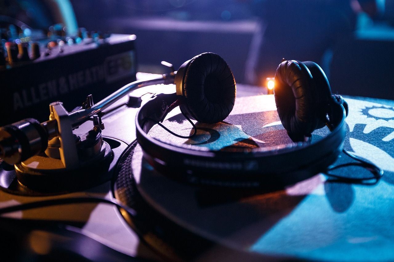 Headphones and a vinyl record on a dj's turntable lit by blue stage lights, with sound mixer equipment in the background.