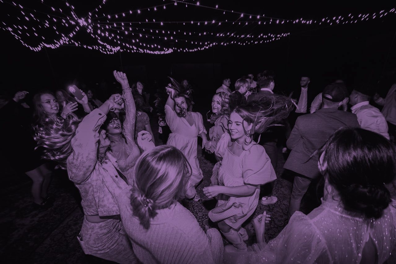 A lively outdoor nighttime party with people dancing under string lights, captured in a monochrome purple hue.